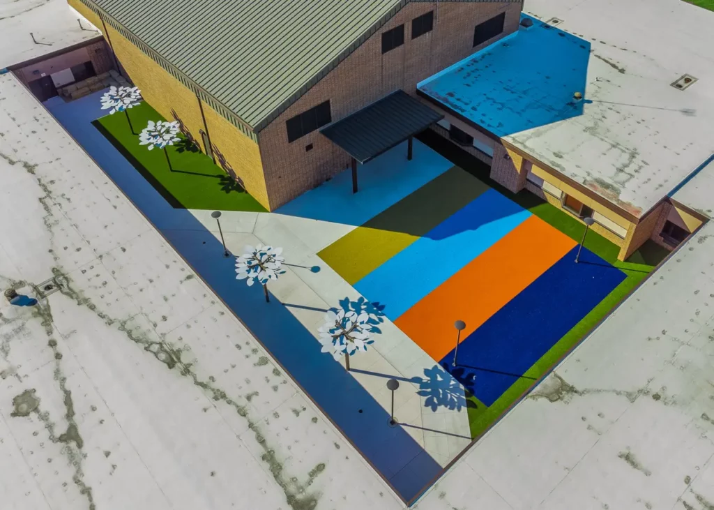 Aerial view of elementary school playground renovation featuring colorful rubber surface courts, artificial turf, and decorative metal flower sculptures.
