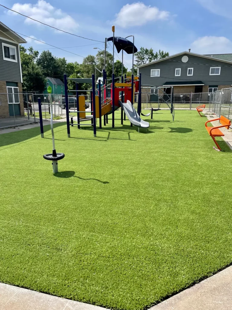 Bright artificial turf playground at Washington Gardens with colorful play structure, slide, spinner, and orange benches, fenced urban setting.