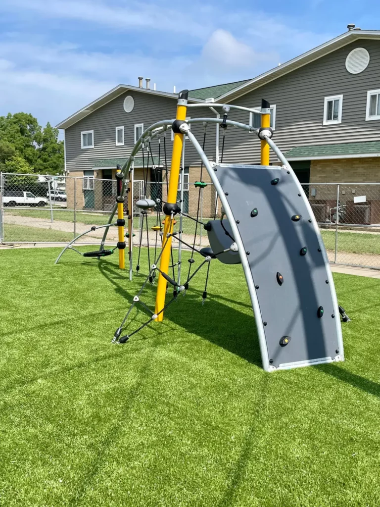 Modern playground climbing structure with rope net and rock wall on lush artificial turf at Washington Gardens municipal park.