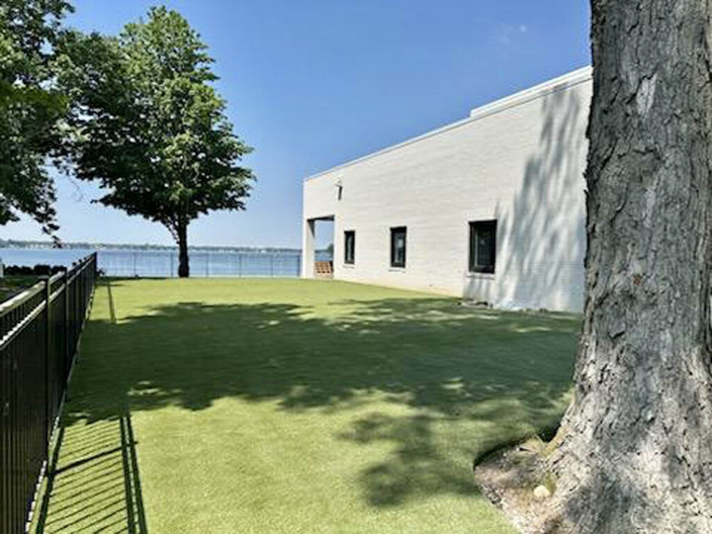 3,000 sq ft K9Grass Elite artificial turf pet yard beside modern lakefront home with black metal fencing and mature trees.