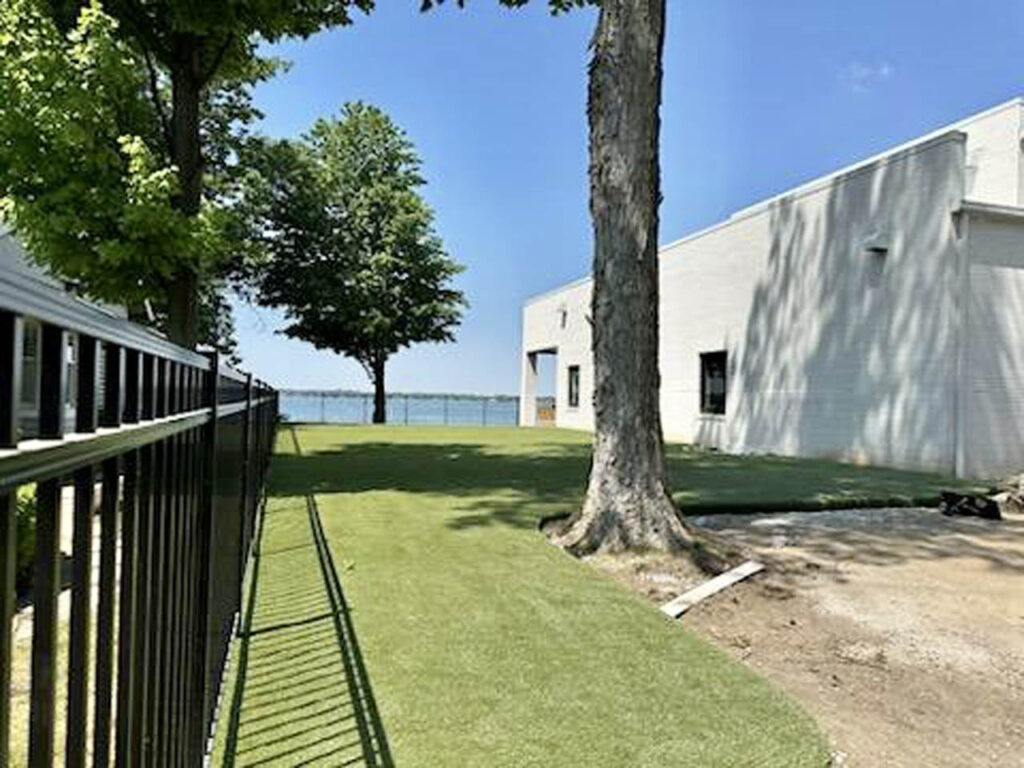 3,000 sq ft K9Grass Elite artificial turf pet yard beside lakefront home with black aluminum fencing and mature trees.