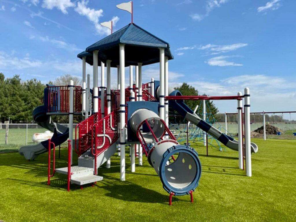 Colorful red, white, and blue playground structure with slides and tube tunnel installed on lush artificial turf at Hoagland Park.