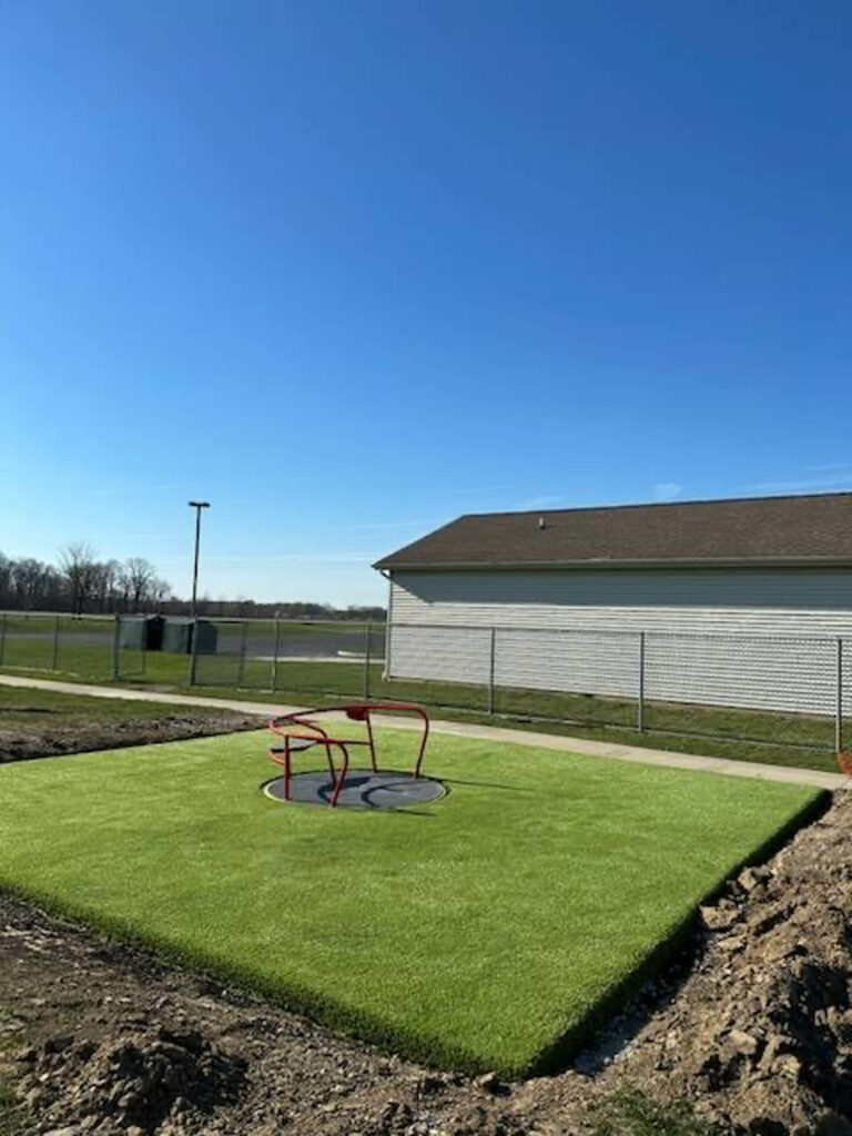 Bright green artificial turf playground area with red spinning seat, adjacent to school building and chain-link fence, under clear blue sky.