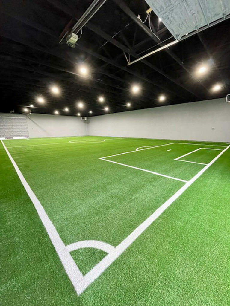 Indoor 6,000 sq ft artificial turf soccer field with white boundary lines, corner arc, and penalty box under bright overhead lighting.