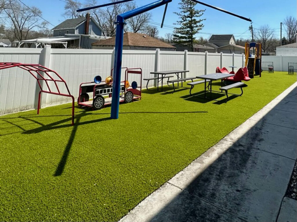 Childcare center playground with artificial turf, red climbing structures, fire truck toy, picnic tables, and white vinyl privacy fence.