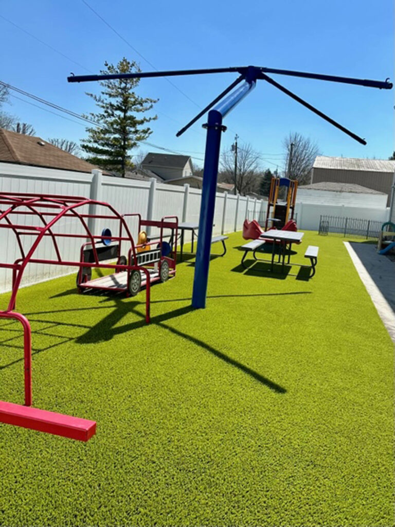 Bright green artificial turf playground at childcare center with red climbing structures, blue spinner pole, and picnic tables.