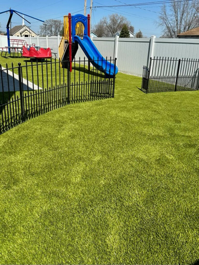 Bright artificial turf playground at childcare center with colorful slide set and black iron safety fencing, white privacy fence backdrop.