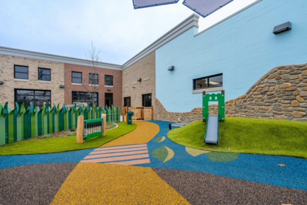 Colorful rubber safety surfacing and artificial turf playground at nonprofit facility with slide, musical instruments, and decorative fencing.