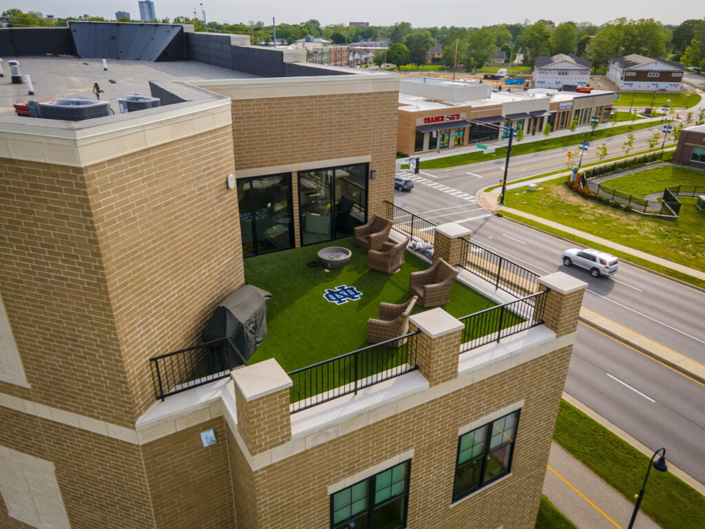 Rooftop terrace with 690 sq ft SportsGrass turf, Notre Dame logo, wicker seating, fire pit, and brick railing overlooking urban street.