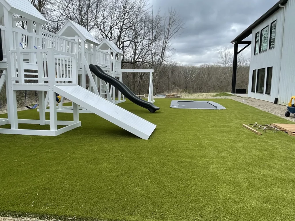 White vinyl playset with black slide and in-ground trampoline on lush artificial turf beside modern farmhouse in Veedersburg, IN.