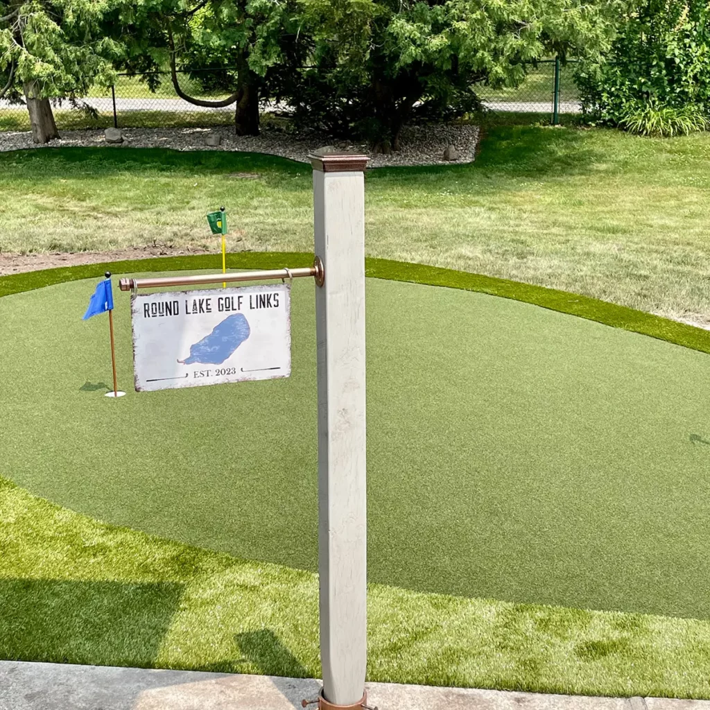 Custom backyard putting green with two holes, flags, and Round Lake Golf Links Est. 2023 sign, surrounded by natural lawn and trees.