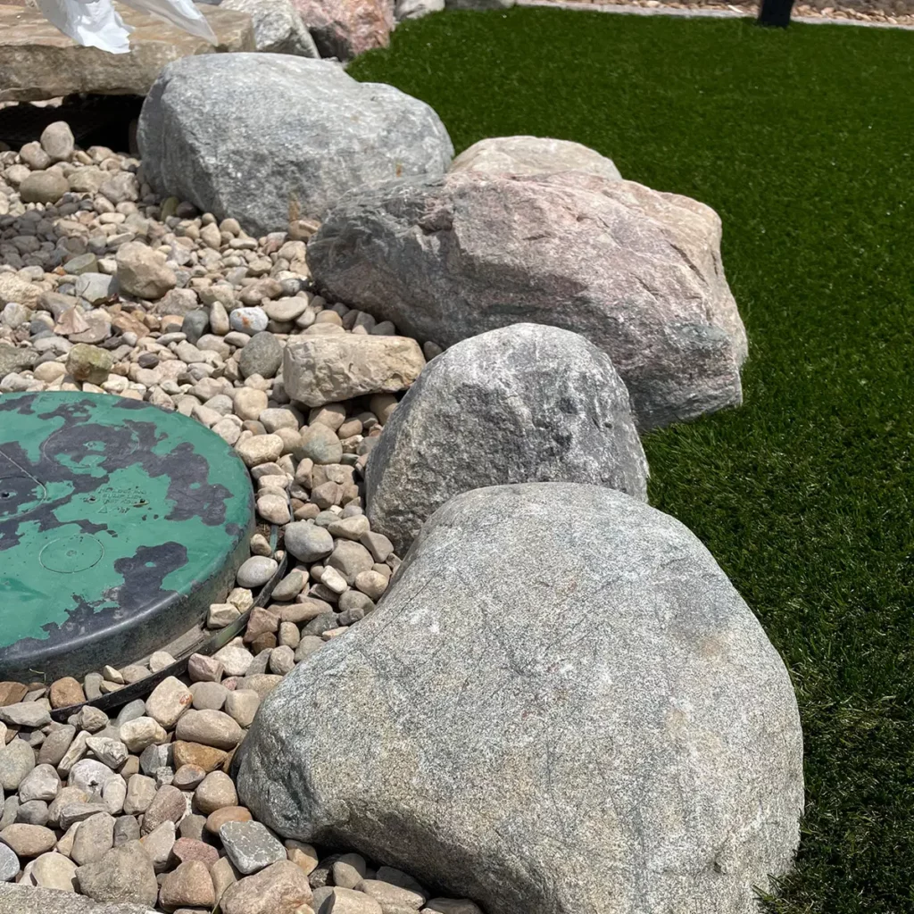 Decorative granite boulders and river rock border artificial turf backyard putting green during residential landscaping installation.
