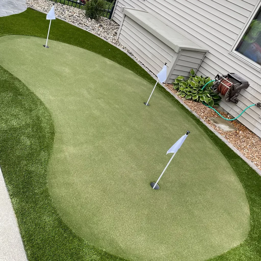 Backyard artificial grass putting green with 3 white flags and holes, bordered by decorative rock and home siding.