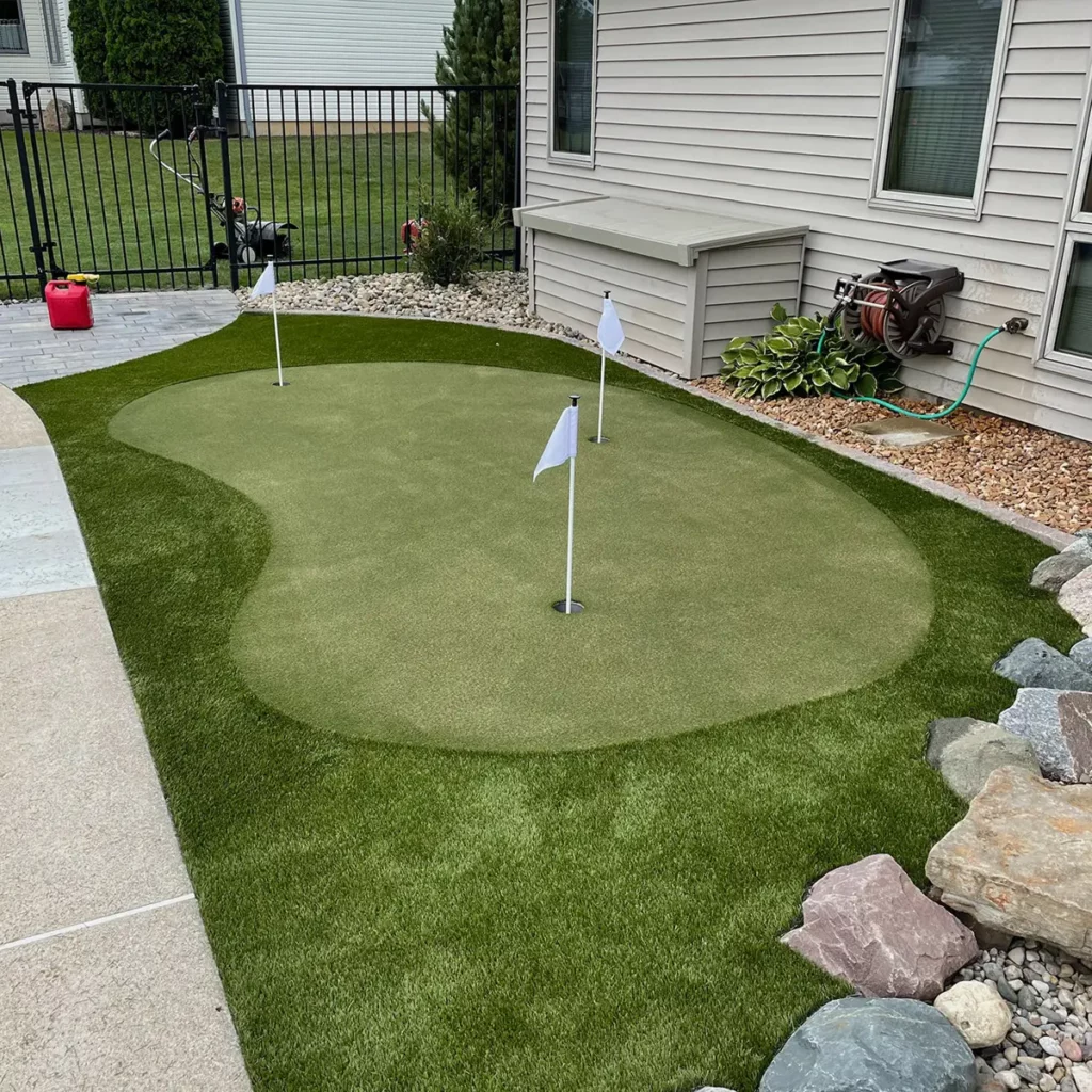 Backyard artificial grass putting green with 3 holes and white flags, bordered by decorative rocks and gravel beside a home.