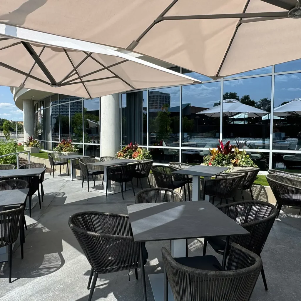 Modern outdoor patio with woven rope chairs, dark tables, large shade umbrellas, tropical planters, and floor-to-ceiling glass facade.