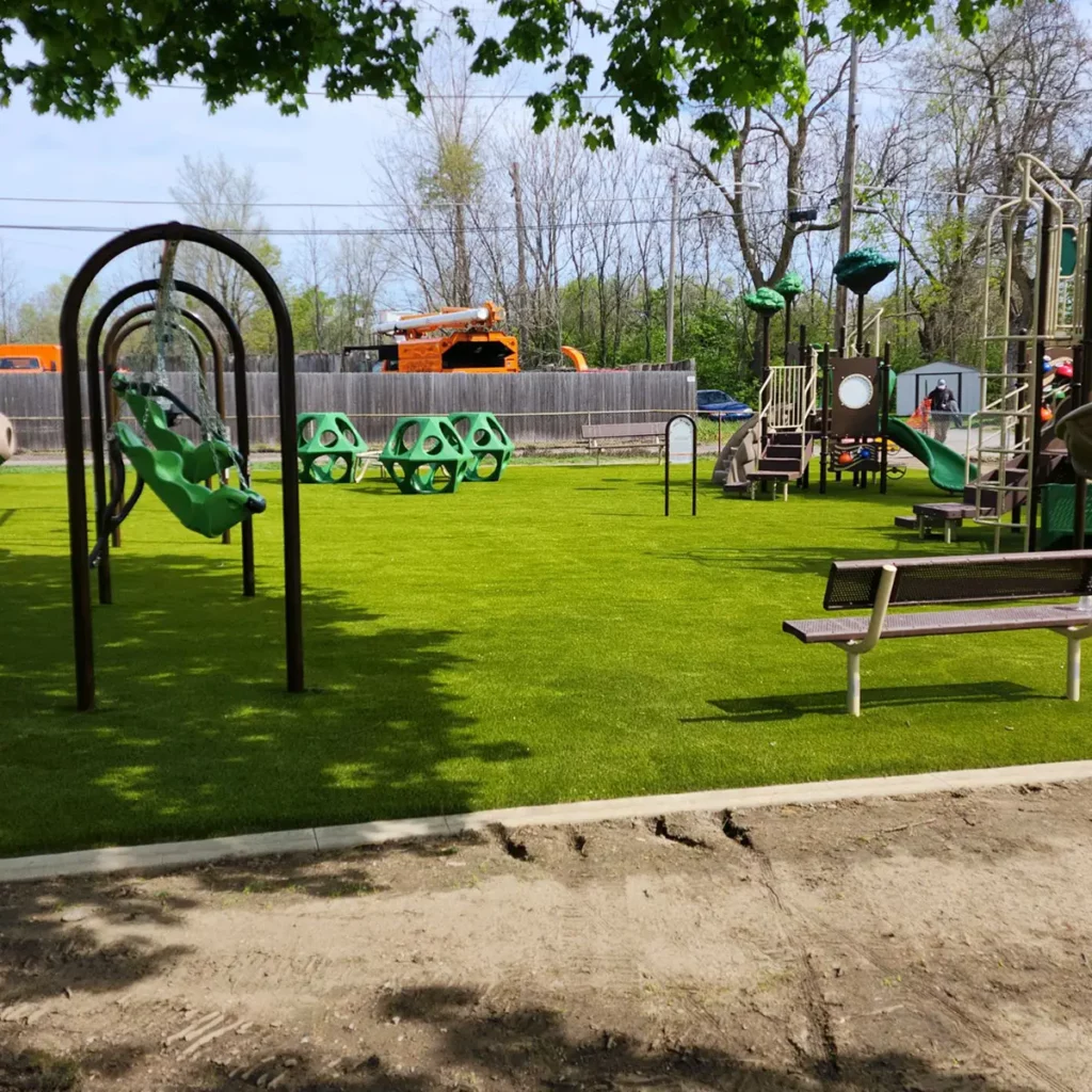 Artificial turf playground in Fort Wayne municipal park with swings, climbing structure, green seating, and bench on bright synthetic grass.