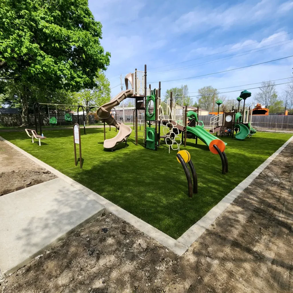 Newly installed artificial turf playground in Fort Wayne municipal park with colorful play structures, slides, and concrete border edging.