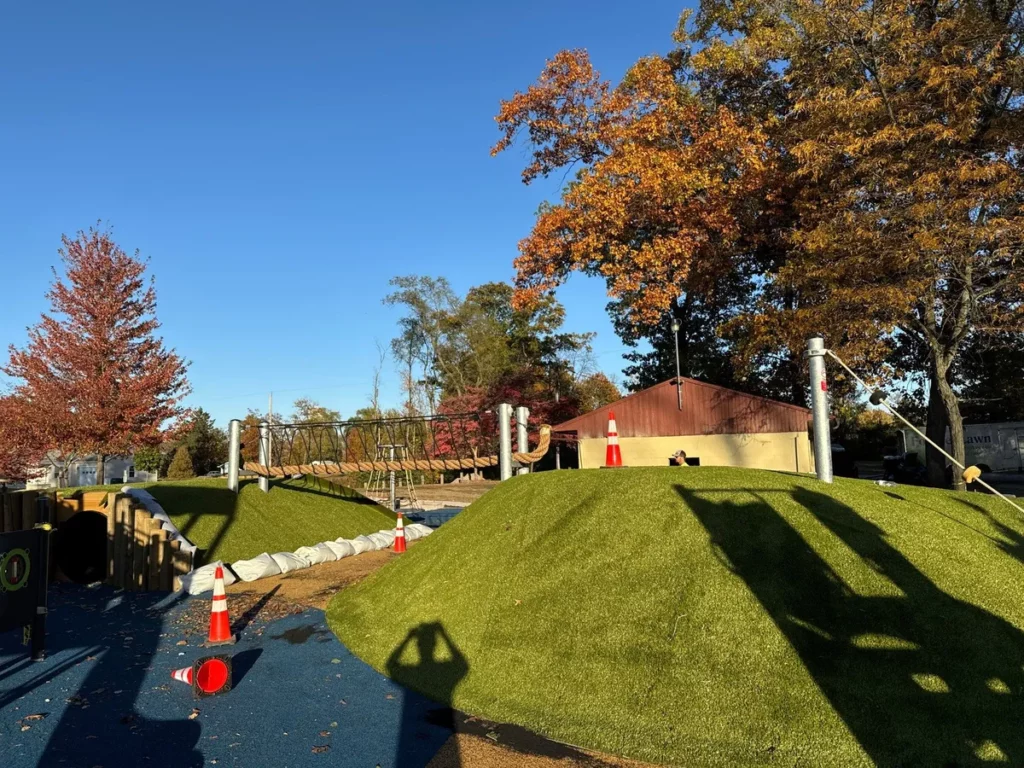Artificial turf hills and rope bridge at North Judson municipal playground under construction, autumn trees, orange safety cones visible.