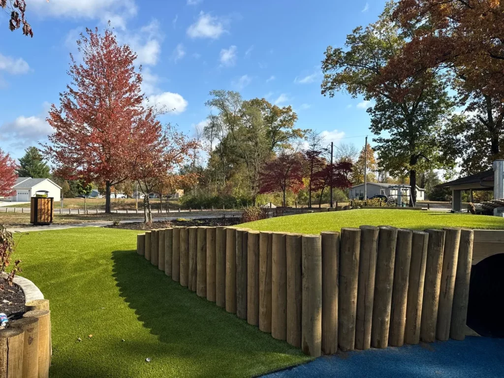 Lush artificial turf playground with timber log retaining wall, rolling hills, and colorful autumn trees at North Judson municipal park.