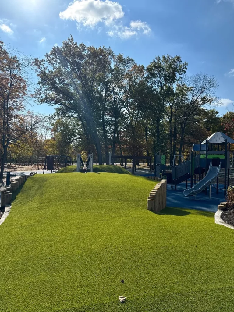 Lush artificial turf playground area with rolling hills, timber borders, and modern play structure with slides, surrounded by autumn trees.