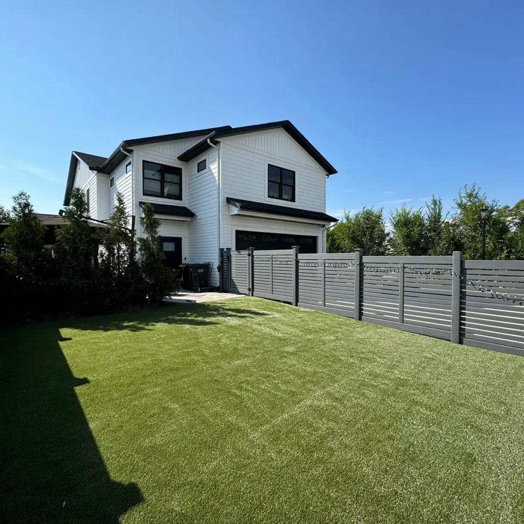 K9Grass Classic+ artificial turf installed in spacious backyard beside modern white farmhouse with gray horizontal privacy fence.