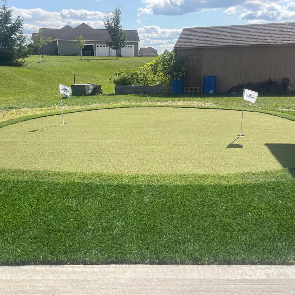 Backyard artificial putting green with two GolfGreens flags, oval shape, surrounded by natural grass, residential neighborhood setting.