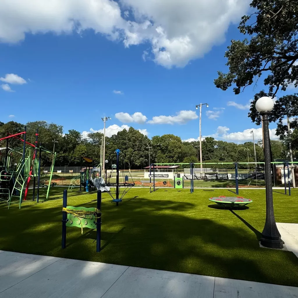 Newly installed Playground Grass Ultra artificial turf at Wabash Park municipal playground with colorful equipment, lamp post, and baseball field beyond.