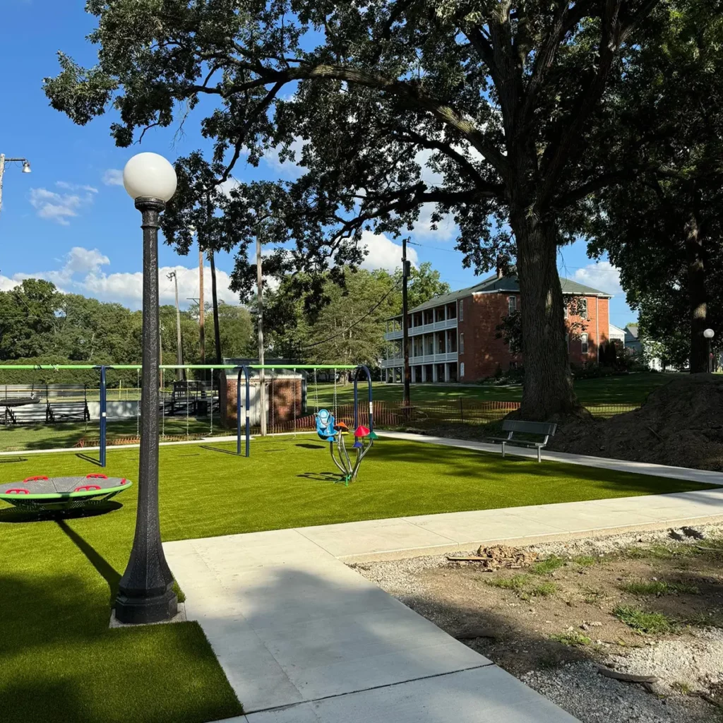 Newly installed bright green artificial turf playground at Wabash Park with swings, spinner, lamp post, and brick building backdrop.