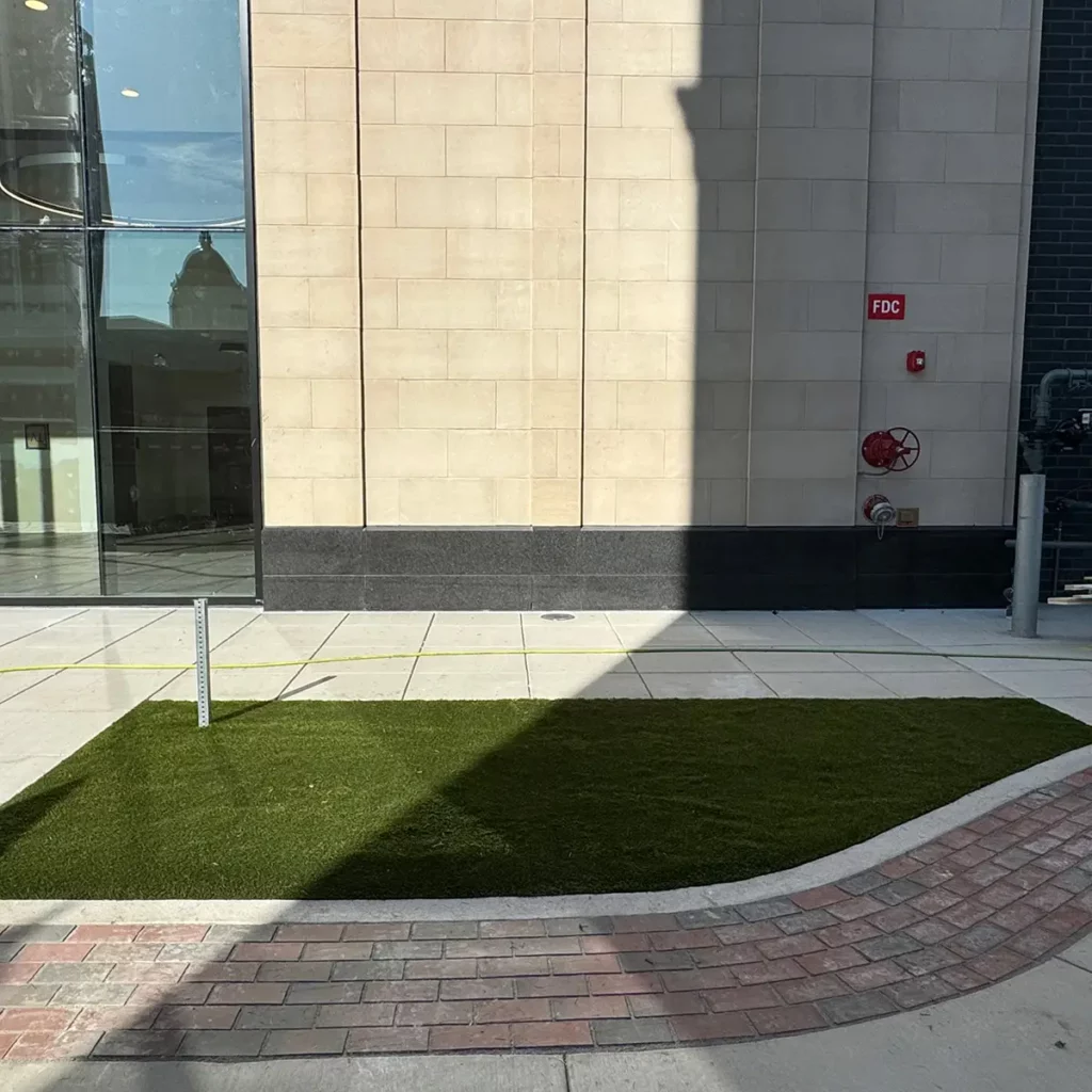 K9Grass Classic+ artificial turf patch installed beside Pearl Event Center entrance, bordered by curved brick pavers and concrete walkway.