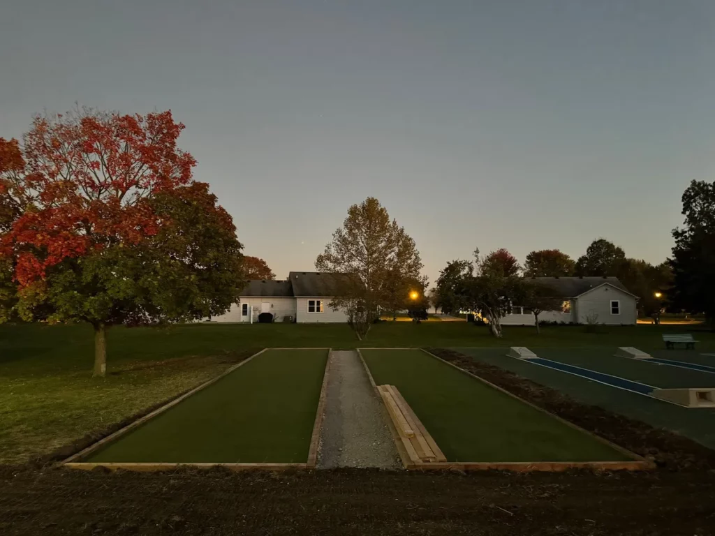 Two newly installed bocce ball courts with artificial turf and gravel center path at dusk, residential New England setting, fall foliage