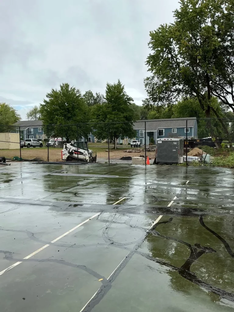 Wet asphalt parking lot with Bobcat skid steer and chain-link fencing during K9Grass dog park installation, South Bend, IN.