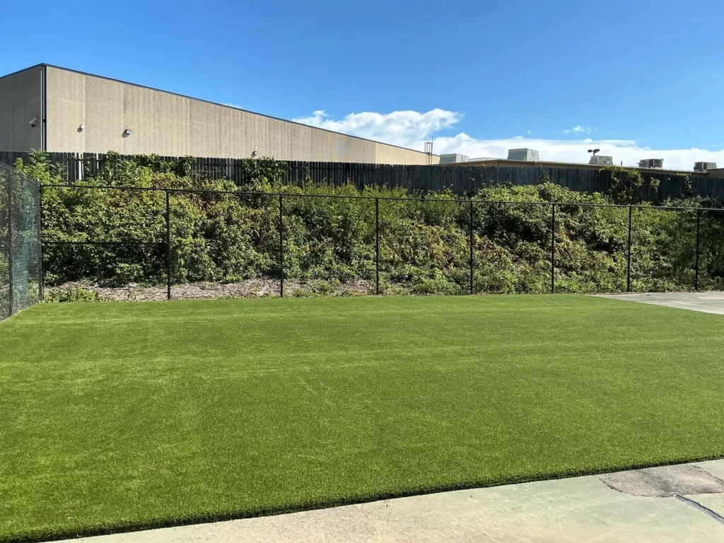 K9Grass artificial turf dog park with black chain-link fence, lush shrubs, and commercial building backdrop in South Bend, Indiana.