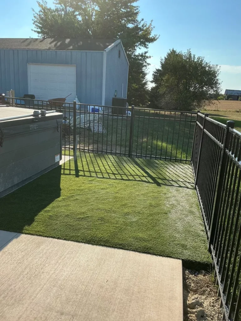 K9Grass Classic+ artificial turf installed in fenced residential dog area beside hot tub, Fort Wayne home with metal barn behind.