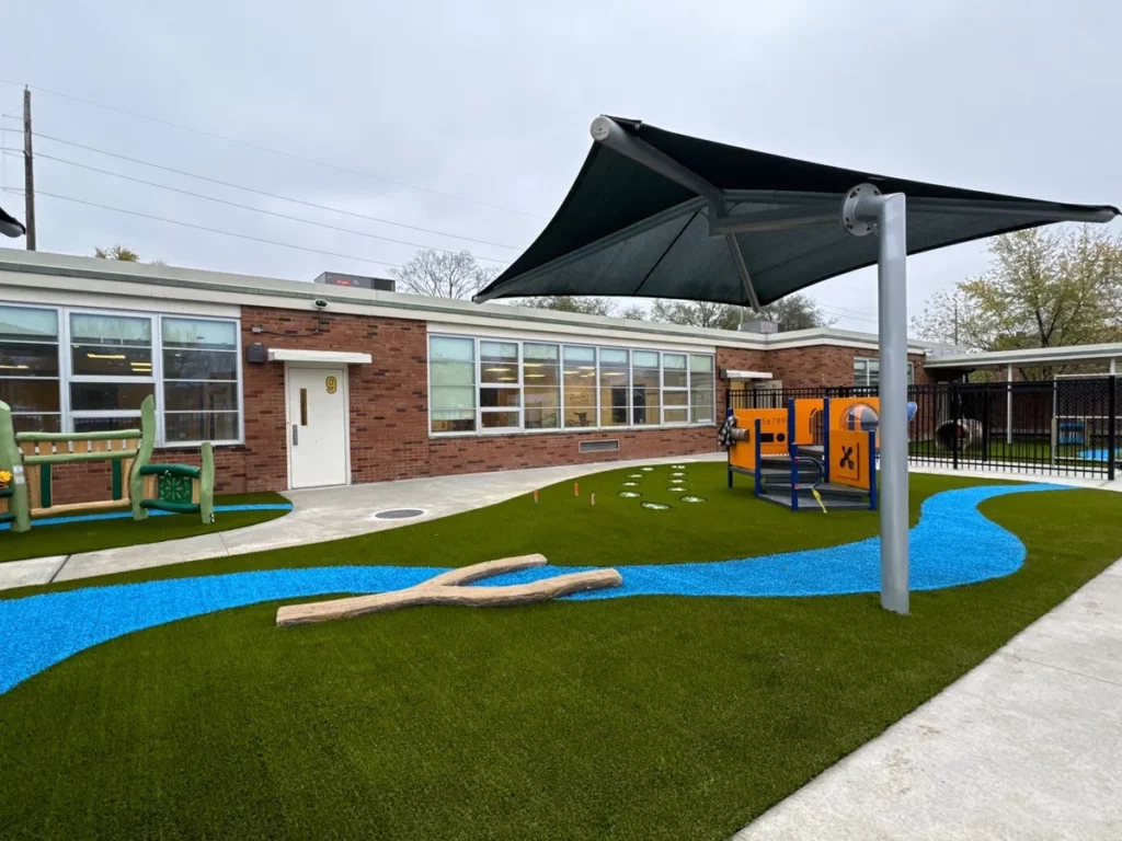 Columbus school playground with synthetic turf, blue rubber river path, shade sail, orange play structure, and brick building backdrop.