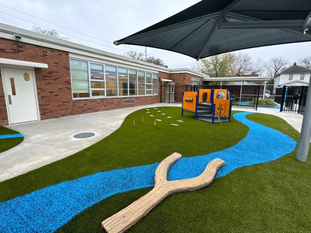 Columbus school playground with blue rubber river path, synthetic turf, orange play structure, shade sail, and Y-shaped log feature.