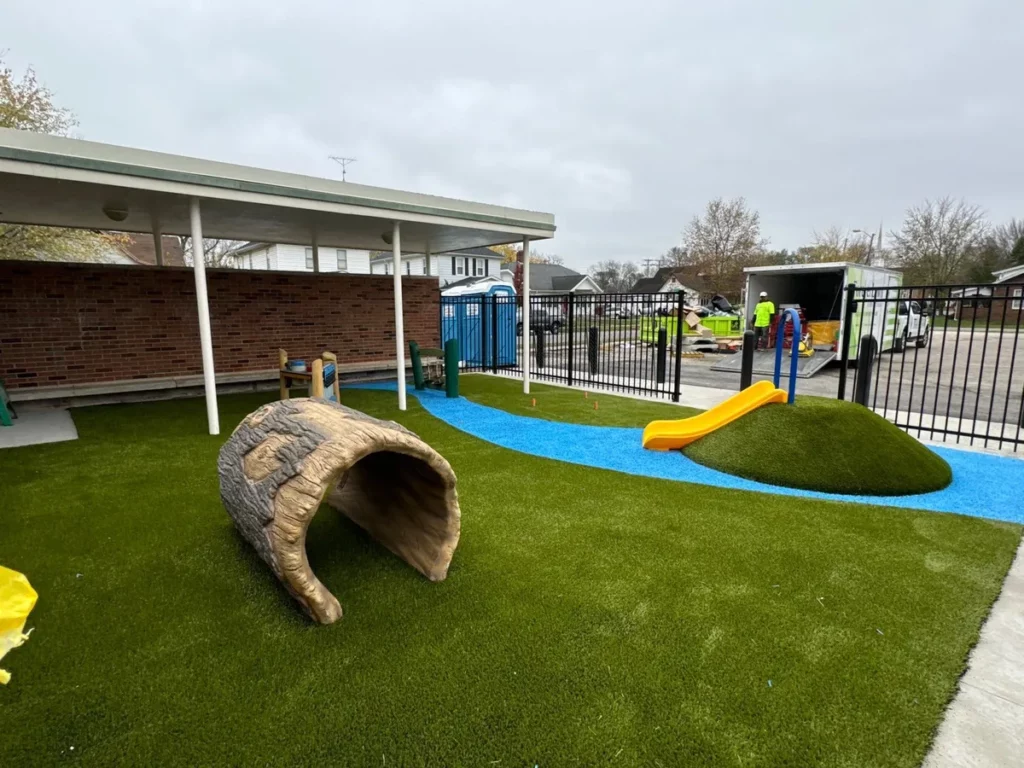 Columbus school playground featuring synthetic turf, blue rubber pathway, log tunnel, grass hill with yellow slide, and covered play area.