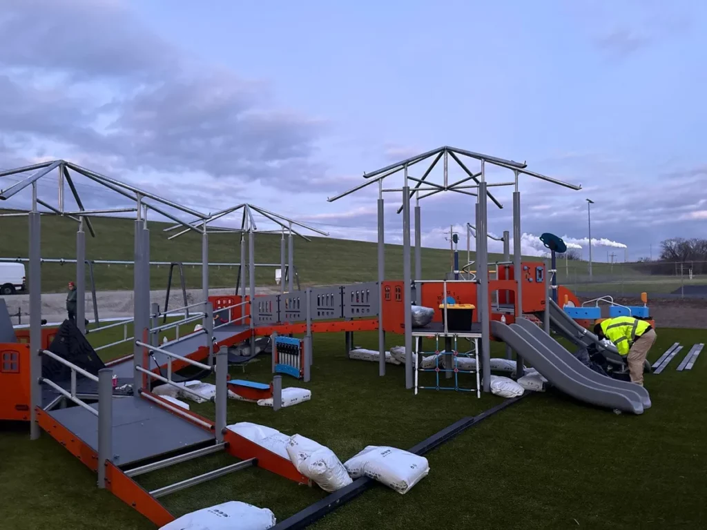 Workers installing orange train-themed playground equipment on artificial turf at elementary school, rooftop frames and slides visible.