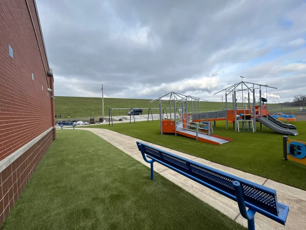 New elementary school playground with Playground Grass Ultra artificial turf, blue benches, orange play structure, and slides beside brick building.