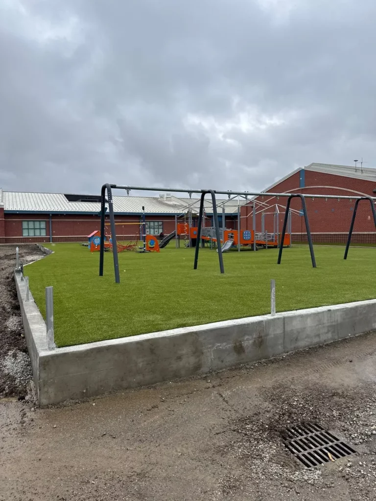 New elementary school playground with artificial turf, swing sets, and colorful play structures surrounded by concrete edging and brick school building.