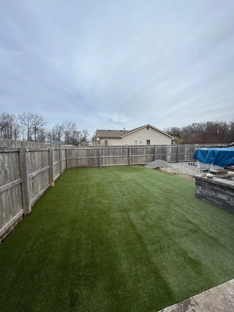 K9Grass Elite artificial turf installed in fenced residential backyard with stone retaining wall and patio edging.