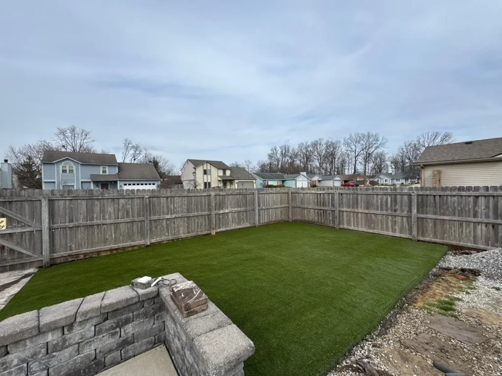 Fenced suburban backyard with K9Grass Elite artificial turf installation, stone retaining wall, gravel border, overcast sky.