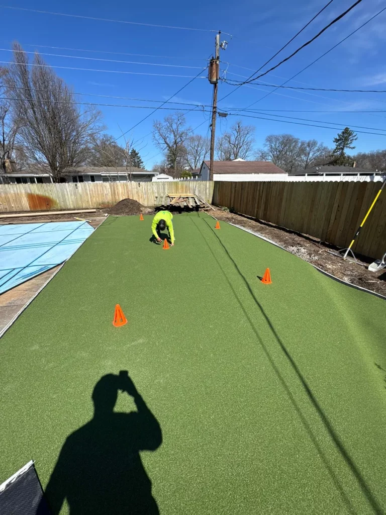 Contractor installs GolfGreens synthetic putting green in fenced Indiana backyard with orange marker cones on bright turf.
