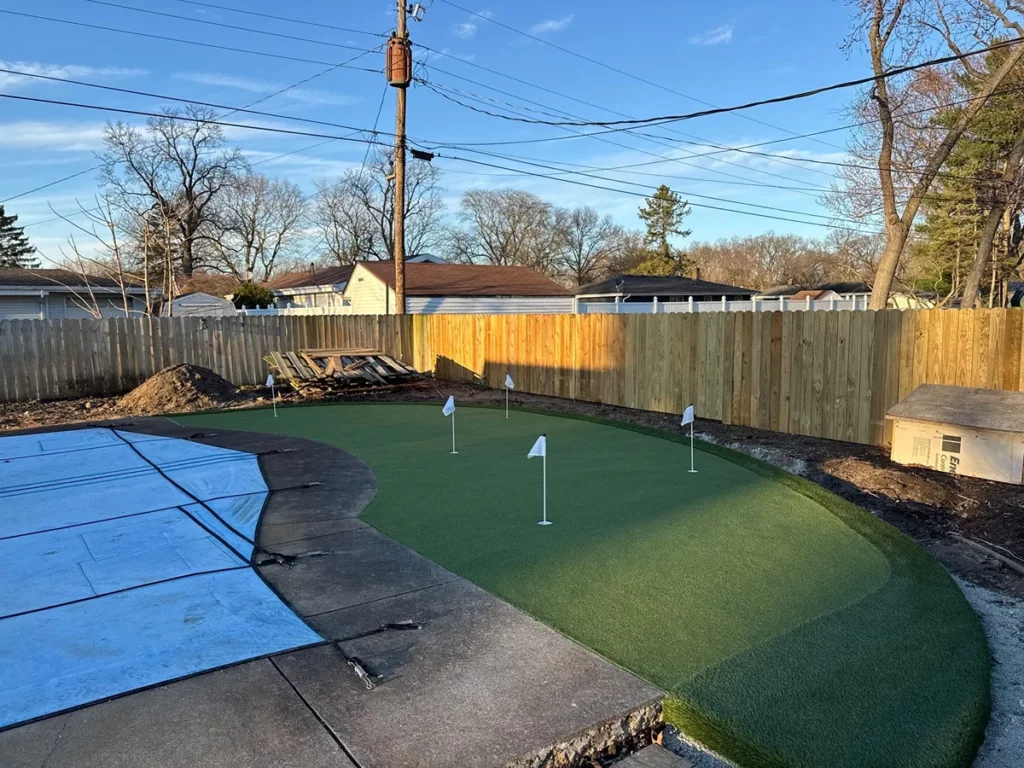 Backyard synthetic putting green with 3 flag holes, curved turf layout, wood privacy fence, adjacent concrete patio, residential setting.