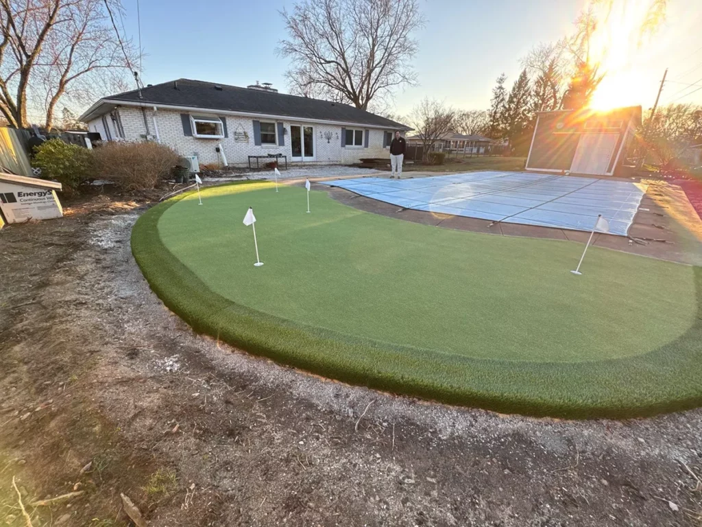 Backyard synthetic putting green with 4 cup flags beside covered pool, ranch home backdrop, sunset lighting, Northeast Indiana