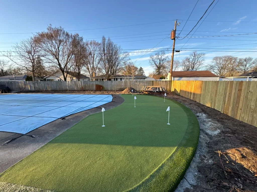 Custom GolfGreens backyard putting green with 4 holes beside covered pool, wood privacy fence, suburban Indiana setting.