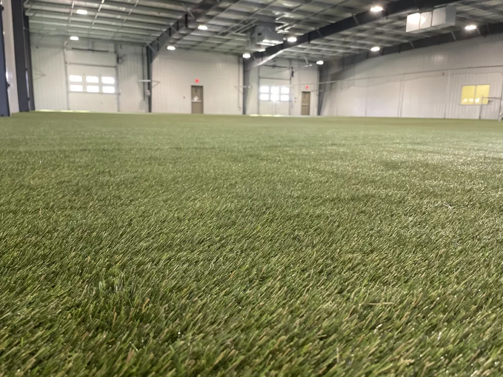 Indoor pet facility with K9Grass artificial turf covering large warehouse space, bright LED lighting, garage doors in background.