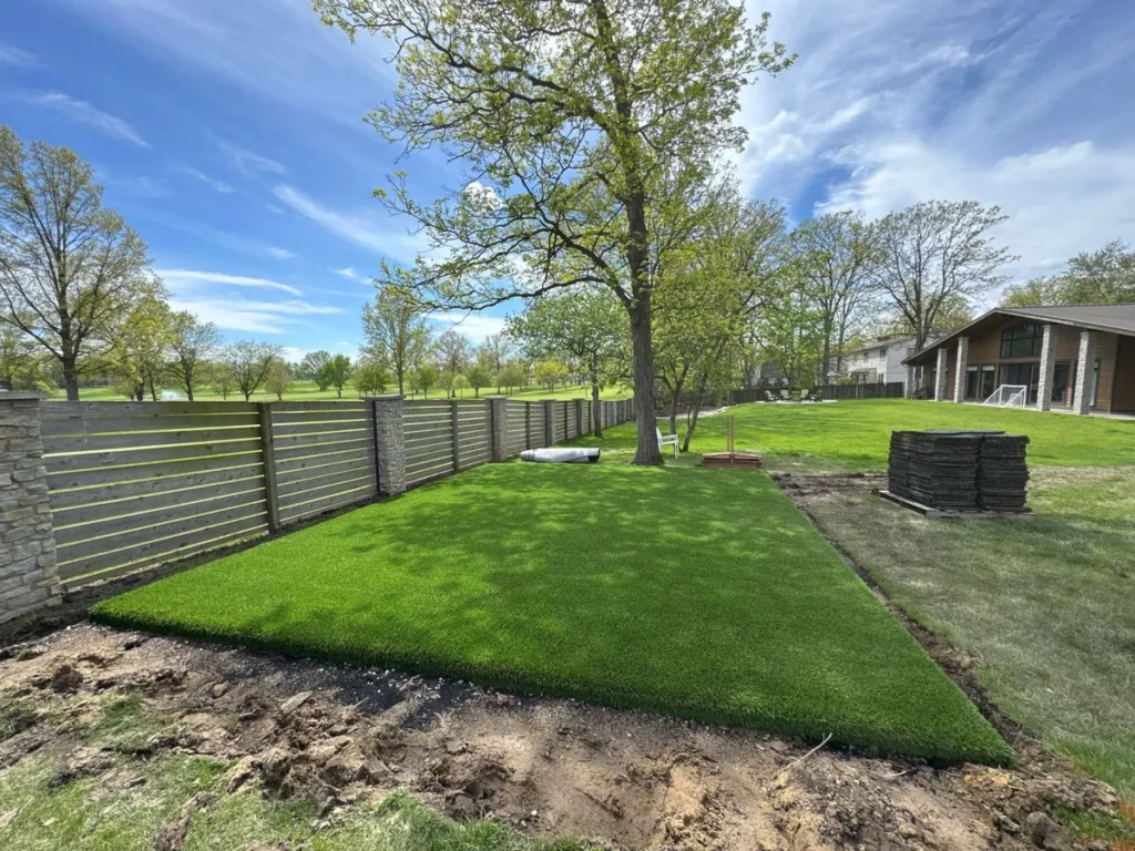 Playground Grass Ultra artificial turf being installed in suburban backyard near modern home, with stacked sod rolls and horizontal fence.