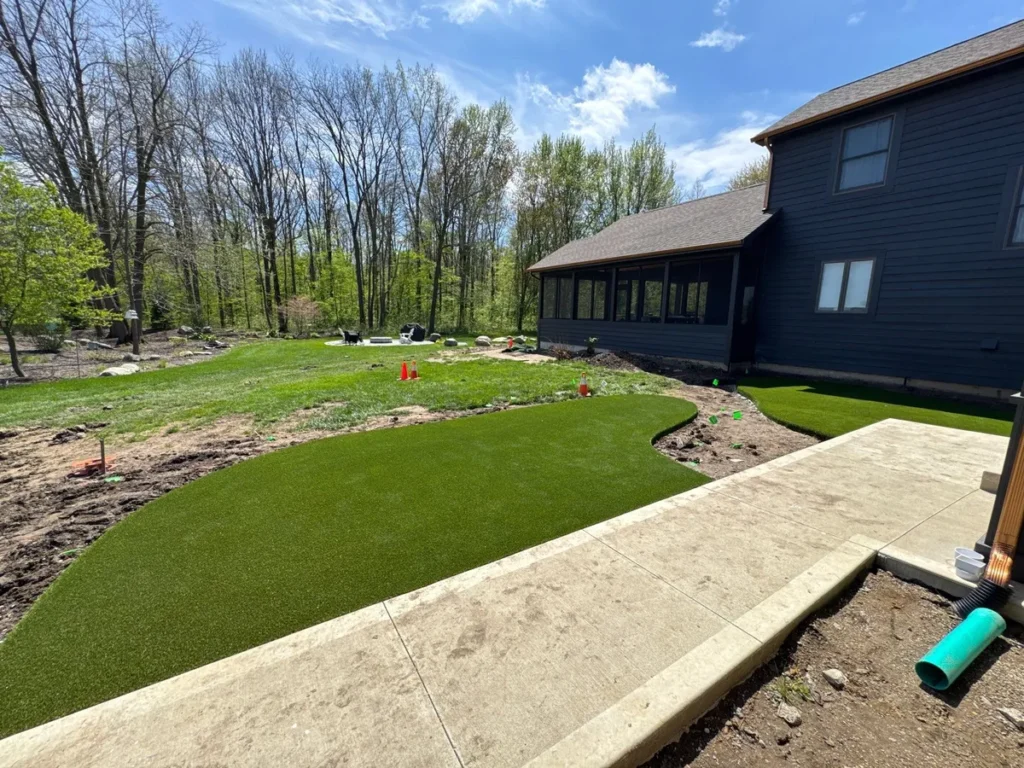 K9Grass artificial pet turf installed beside concrete patio of dark blue two-story home with wooded backyard, Indiana