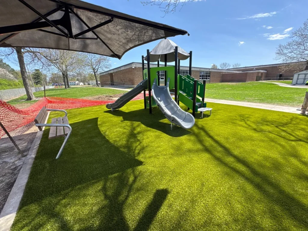 Synthetic turf playground with dual slides, shade canopy, and bench at school; red safety fencing marks nearby construction zone.