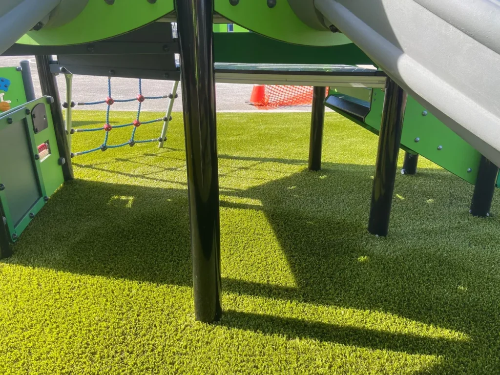 Bright green artificial turf installed under a colorful school playground structure with slides and rope climbing net in Fort Wayne.